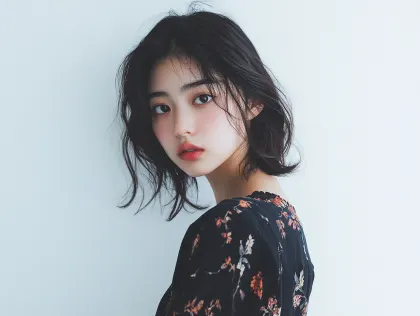 ショートヘアの女性の優しい視線 自然体ポートレート。清潔感のある白い背景の前で、黒い花柄ワンピースを着たショートヘアの若い女性が、柔らかな眼差しでカメラを見つめる美しいポートレート写真です。