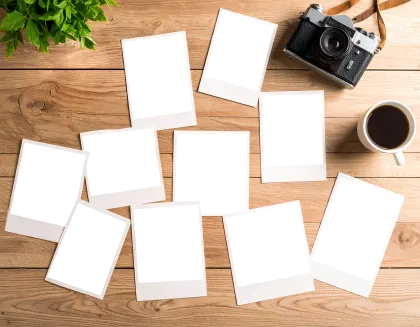 木製デスクに散らばるインスタント写真フレームとレトロカメラ、コーヒーの俯瞰。温かみのある木製デスクに、空白のインスタント写真フレームが複数散らばり、アンティークカメラと温かいコーヒーが置かれた真俯瞰の風景です。